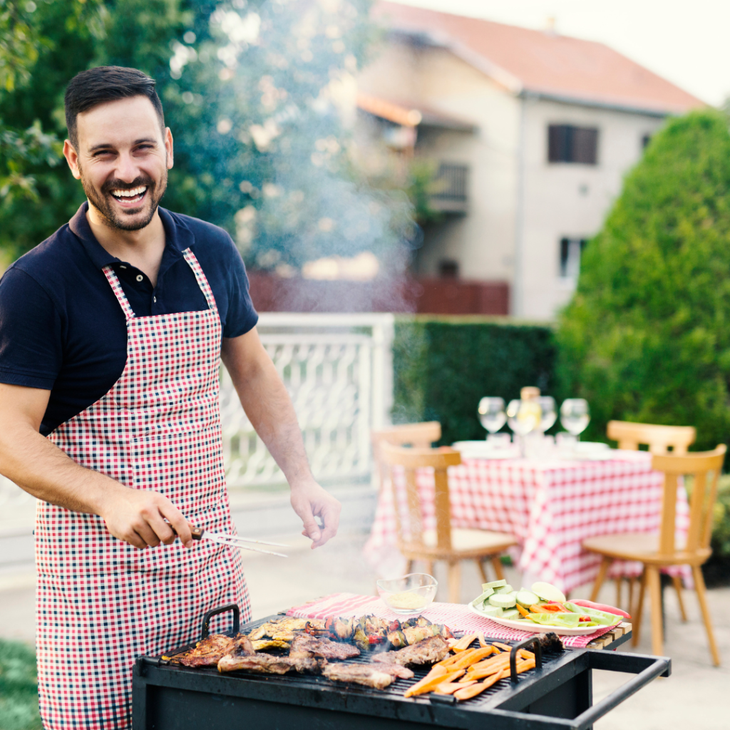 Man grilling in his backyard, different assortments of meats and veggies on the grill with a table set up in the background 